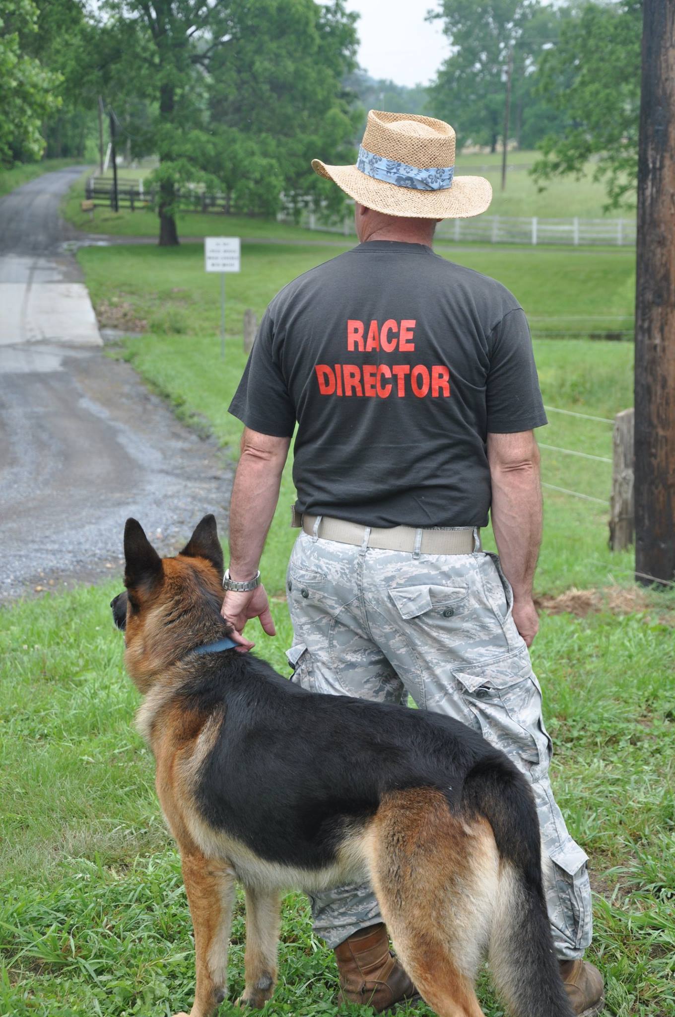 Ray Waldron back to the camera. He is wearing a shirt that says Race Director. He is holding
        the collar of his German Shepard dog while they both look off in the distance.
